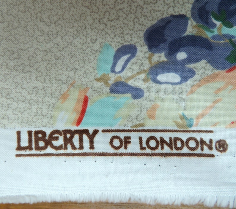 May include: Close-up of Liberty of London fabric. The fabric displays a floral pattern with blue, teal, and peach flowers on a beige backdrop. The brand name is printed in brown letters.