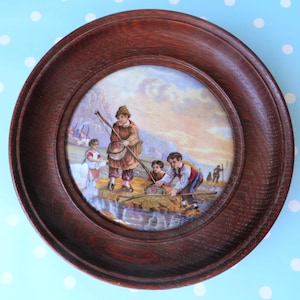 May include: A circular dark brown wooden frame surrounds a central ceramic tile featuring a colourful illustration of three figures fishing in a stream. The scene depicts individuals in period clothing using nets and poles near a grassy bank.
