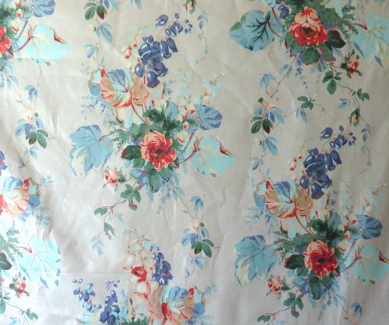 May include: A floral fabric with a pale beige backdrop. The pattern showcases clusters of red roses, blue and pale blue leaves, and purple flowers. The design is a classic, vintage style.