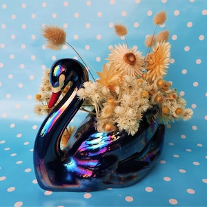 May include: A blue iridescent ceramic swan vase with dried flowers. The vase is decorated with a white polka dot pattern.