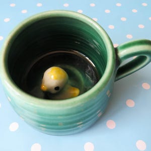 May include: A ceramic mug with a glossy green glaze and horizontal ribbed texture. The interior of the mug features a small, three-dimensional ceramic duckling figure with a white body, yellow head, and black eyes attached to the bottom.