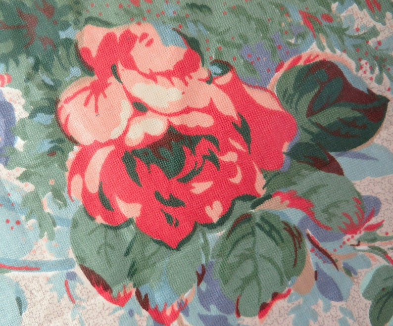 May include: Close-up of a floral fabric featuring a large, vibrant red rose with pink and peach accents. The rose is surrounded by green leaves and a background of light blue and beige tones, creating a vintage aesthetic.