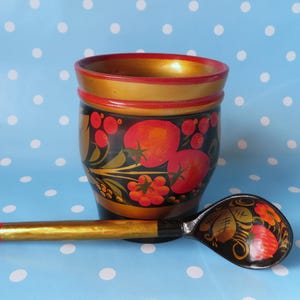 May include: A hand-painted wooden cup and spoon set. The cup is black with red and orange floral designs, and a gold rim. The spoon has a similar design. The background is a light blue with white polka dots.