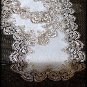 May include: A white table runner with a delicate lace design in a beige color. The lace features intricate floral patterns and scalloped edges.