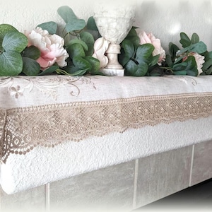 May include: A beige linen fireplace mantel scarf with a delicate lace trim and floral embroidery. The scarf is decorated with a garland of eucalyptus leaves and pink roses.