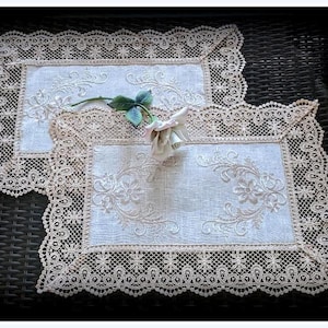 May include: Two delicate white lace doilies with embroidered floral designs. A single white rose with green leaves rests on top of the doilies.