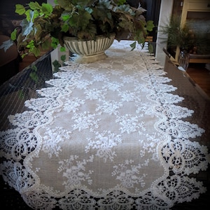 May include: Cream-colored lace table runner with a floral pattern and scalloped edges. The runner is placed on a dark surface, with a decorative vase of greenery in the background. The lace design adds a touch of elegance.