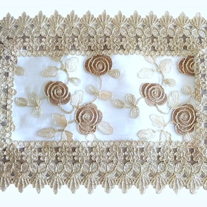 May include: A white table runner with a gold lace border and brown floral embroidery.