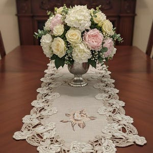 May include: A dining table centerpiece featuring a silver vase overflowing with white, pink, and yellow flowers. A cream-colored lace runner with floral embroidery and a scalloped edge sits beneath the vase, adding an elegant touch to the setting.