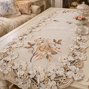 May include: Beige table runner with embroidered floral design. The runner features a large central flower and delicate lace edging. The color palette includes beige, cream, and brown tones, creating a vintage aesthetic. The runner is placed on a white table.