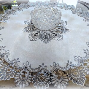May include: A white round table runner with a silver lace design. The runner has a scalloped edge and a large floral design in the center. A clear glass bowl is sitting on top of the runner.