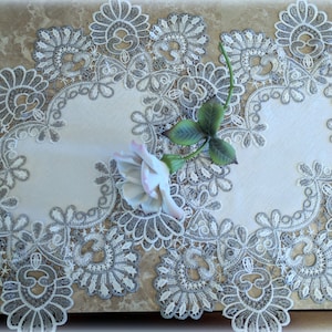 May include: A white table runner with a silver lace border. The lace features a delicate floral design. A single white rose with green leaves is placed on the runner.