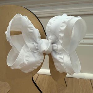 May include: A large, white hair bow with a textured, ruffled design. The bow is made of a thick, satin-like material and is displayed on a brown, cutout stand. The bow is centered and symmetrical, with a prominent knot in the middle.
