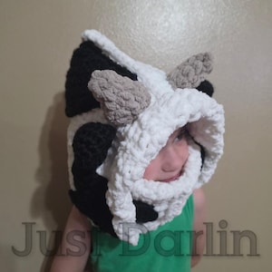 May include: A plush, chunky crochet hood featuring a black and white cow-inspired pattern. The hood includes two small, textured light brown horns on top and a thick, soft white border framing the face opening. The text Just Darlin is visible.