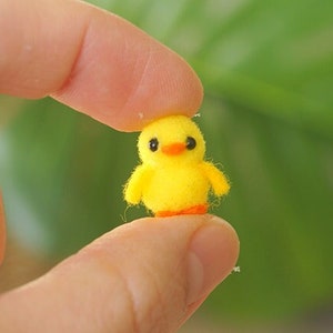 May include: A small yellow felt chick with black eyes and an orange beak. The chick is sitting on a finger.