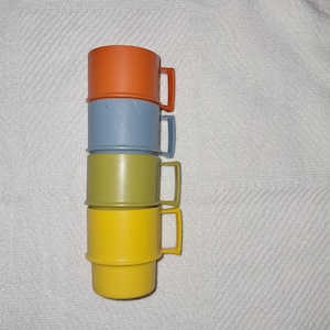 Könnte beinhalten: Ein Stapel von vier bunten Plastikbechern. Die Becher sind orange, blau, grün und gelb. Jeder Becher hat einen Griff. Die Becher sind übereinander gestapelt.