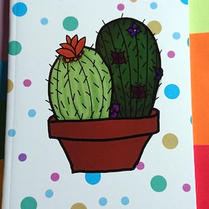 May include: A white notebook with a colorful polka dot pattern. The notebook features a drawing of two cacti in a brown pot with purple flowers.