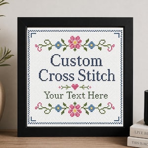 May include: A framed cross stitch sampler featuring the text Custom Cross Stitch and Your Text Here in dark blue lettering. The design is decorated with pink and blue floral motifs, green vines, and a small pink heart centered between the lines.