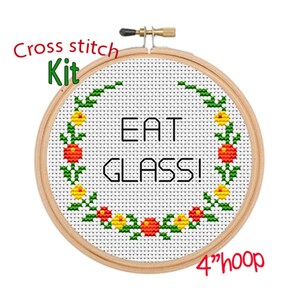 Può includere: Kit di punto croce con un cerchio da 10 cm di diametro. Il tessuto bianco è decorato con una ghirlanda di fiori verdi con fiori rossi e gialli. Il testo "EAT GLASS!" è ricamato con filo nero.