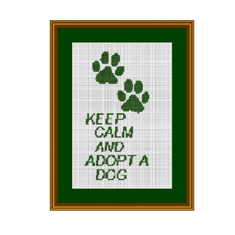 May include: A framed cross-stitch design with a green background and white fabric. The design features two green paw prints at the top and the text "KEEP CALM AND ADOPT A DOG" in green letters.