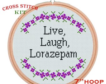 May include: A cross-stitch kit with a 7-inch hoop. The design features the words "Live, Laugh, Lorazepam" in black, surrounded by a border of purple flowers and green leaves. The words "Cross Stitch Kit" are in red and green.