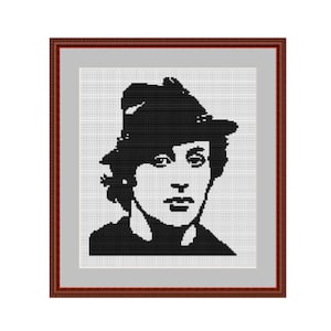 Könnte beinhalten: Schwarzweißes Kreuzstich-Porträt einer Person mit Hut. Die Person hat kurzes, welliges Haar und blickt direkt auf den Betrachter.