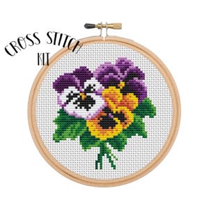 May include: Cross stitch kit featuring a colorful bouquet of three purple, yellow, and orange pansies with green leaves. The kit is displayed in a wooden embroidery hoop.