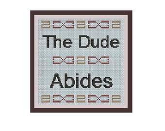 May include: Cross-stitch pattern with a brown frame and a light blue background. The pattern reads "The Dude Abides" with a brown and gold design around the text.