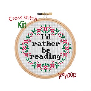 Peut inclure: Kit de point de croix avec un cercle de 18 cm de diamètre et un tissu blanc. Le tissu est brodé d'une couronne de fleurs avec le texte "I'd rather be reading" en noir.