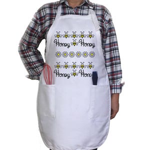 May include: White apron with two pockets and a design of bees and daisies. The text "Honey Honey Honey Home" is printed on the apron.