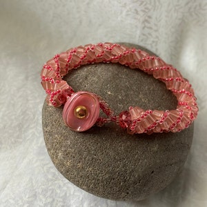 Könnte beinhalten: Ein rosafarbenes Armband mit einem rosafarbenen Knopfverschluss. Das Armband besteht aus kleinen, quadratischen Perlen und ist spiralförmig geflochten.