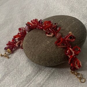 Puede incluir: Una pulsera de cuentas hecha a mano en tonos rojos, rosas y blancos, con un diseño de dragón. La pulsera, con un cierre dorado, está colocada sobre una piedra gris. Las cuentas están dispuestas para formar el dragón y elementos florales.