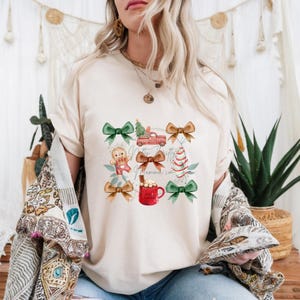 May include: Cream-colored t-shirt featuring a festive design. The graphic includes a red truck carrying a Christmas tree, a gingerbread man, a Christmas tree, a mug of hot cocoa, and bows. The text "Merry Thrifty Momma Co." is also present.