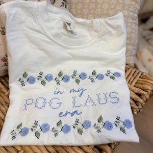 May include: A white short-sleeved t-shirt featuring a cross-stitch style embroidery design. The graphic includes blue floral vines at the top and bottom with the text "in my POG LAUS era" embroidered in blue across the center of the garment.
