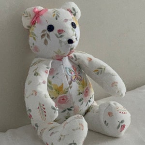 May include: A white teddy bear with a floral print, pink bow, and black button eyes. The bear is sitting on a white surface.