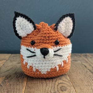 May include: A crocheted fox-shaped pouch with orange and white fur, black eyes, and a black nose. The pouch is sitting on a wooden surface.