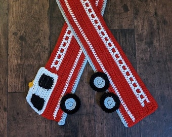 May include: Red and white crocheted scarf with a fire truck design. The scarf has white trim and a gray and black fire truck with wheels.
