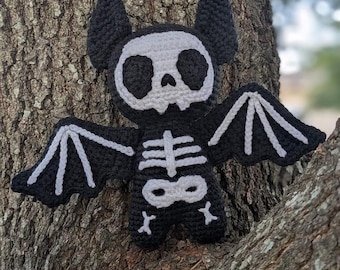 Op de afbeelding: Een zwart-witte gehaakte vleermuis met een schedelgezicht en skeletontwerp. De vleermuis heeft grote oren en uitgespreide vleugels. De vleugels en skeletdetails zijn wit. Een decoratief item met Halloween-thema.