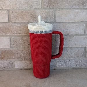 May include: A crocheted red mug with a white lid and a grey stripe. The mug has a handle and is made of yarn.