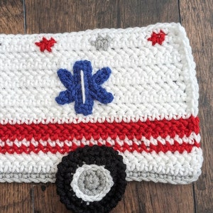 May include: A white crocheted ambulance with red and white stripes, a blue star of life, and a black tire with a gray center.