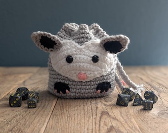 May include: A gray and white crocheted opossum-shaped dice bag with black eyes and a pink nose. The bag is filled with black dice with yellow numbers.