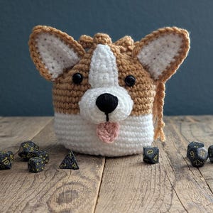 Könnte beinhalten: Ein gehäkelter Würfelbeutel in Form eines Corgis. Die Tasche ist braun und weiß mit schwarzen Augen und einer rosa Zunge. Die Tasche ist gefüllt mit schwarzen Würfeln mit gelben Zahlen.
