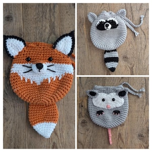 Könnte beinhalten: Drei gehäkelte Tier-Taschen mit Kordelzug. Die Taschen sind ein Fuchs, ein Waschbär und ein Opossum. Die Fuchstasche ist orange und weiß, die Waschbär-Tasche ist grau und schwarz und die Opossum-Tasche ist grau und rosa.