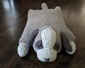 May include: A crocheted dog-shaped pillow with a gray and white color scheme. The pillow is lying on a wooden floor.