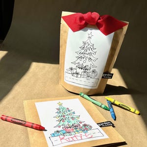 May include: A brown paper bag with a red ribbon and a Christmas tree coloring page. The bag has the text "garden in a bag" on it. There are crayons on a brown paper surface with a Christmas tree coloring page that has the name "Katie" written on it.