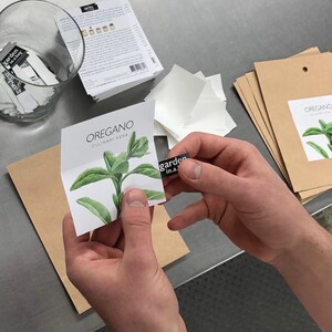 May include: A white card with a watercolor illustration of an oregano plant. The card has the text "OREGANO CULINARY HERB" printed on it. The card is being held by a person's hand, and a small black sticker with the text "garden in.a." is being placed on the card.