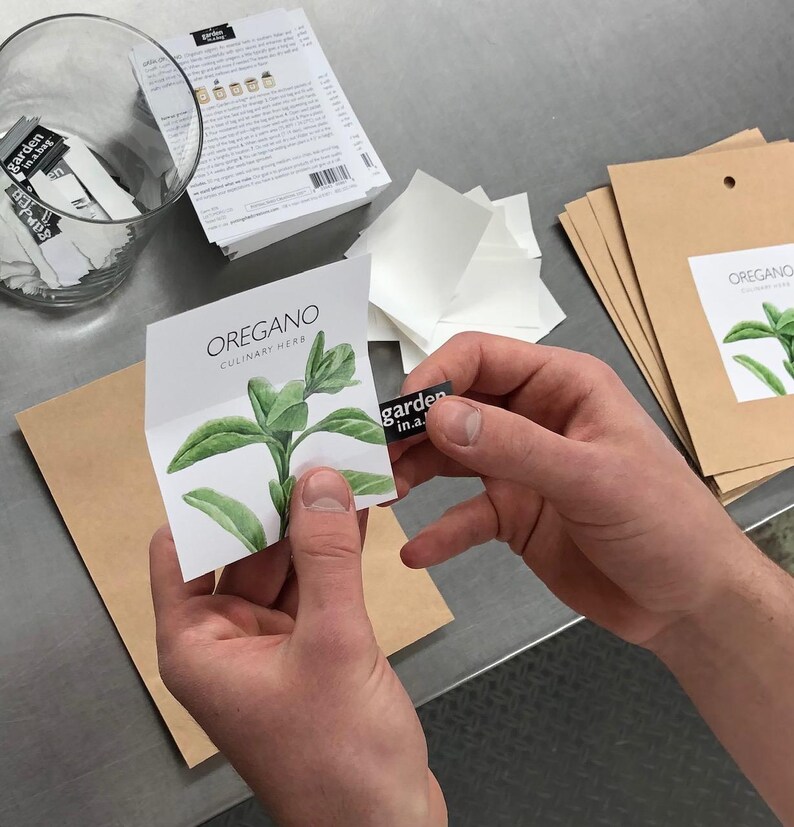 May include: A white card with a watercolor illustration of an oregano plant. The card has the text "OREGANO CULINARY HERB" printed on it. The card is being held by a person's hand, and a small black sticker with the text "garden in.a." is being placed on the card.