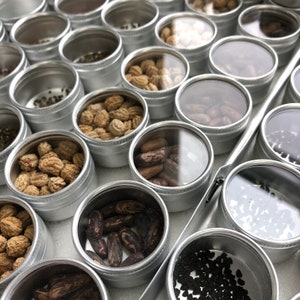 May include: A close-up of a variety of small round containers filled with different types of seeds. The containers are made of metal and have a silver finish. The seeds are arranged in a grid pattern.