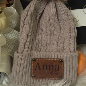 May include: A taupe cable-knit beanie with a brown faux fur pom-pom. The hat features a brown leather patch with the name "Anna" and the word "Elise" below it. The beanie is a winter accessory.
