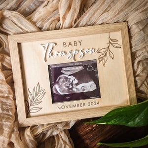 May include: A wooden frame with a light brown background. The frame has a white cutout of the text "Baby Thompson" and a small ultrasound image with the date "November 2024".